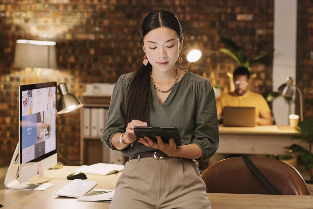 A woman wearing a green shirt, with khaki pants and gold jewelry accessories, is in an dimly lit office holding a tablet in an office leaning on the front of their 90 degree angle desk with an iMac computer to her left. Behind her is a man seated, wearing a yellow collared shirt, is seated behind his desk, in-front of a laptop. 