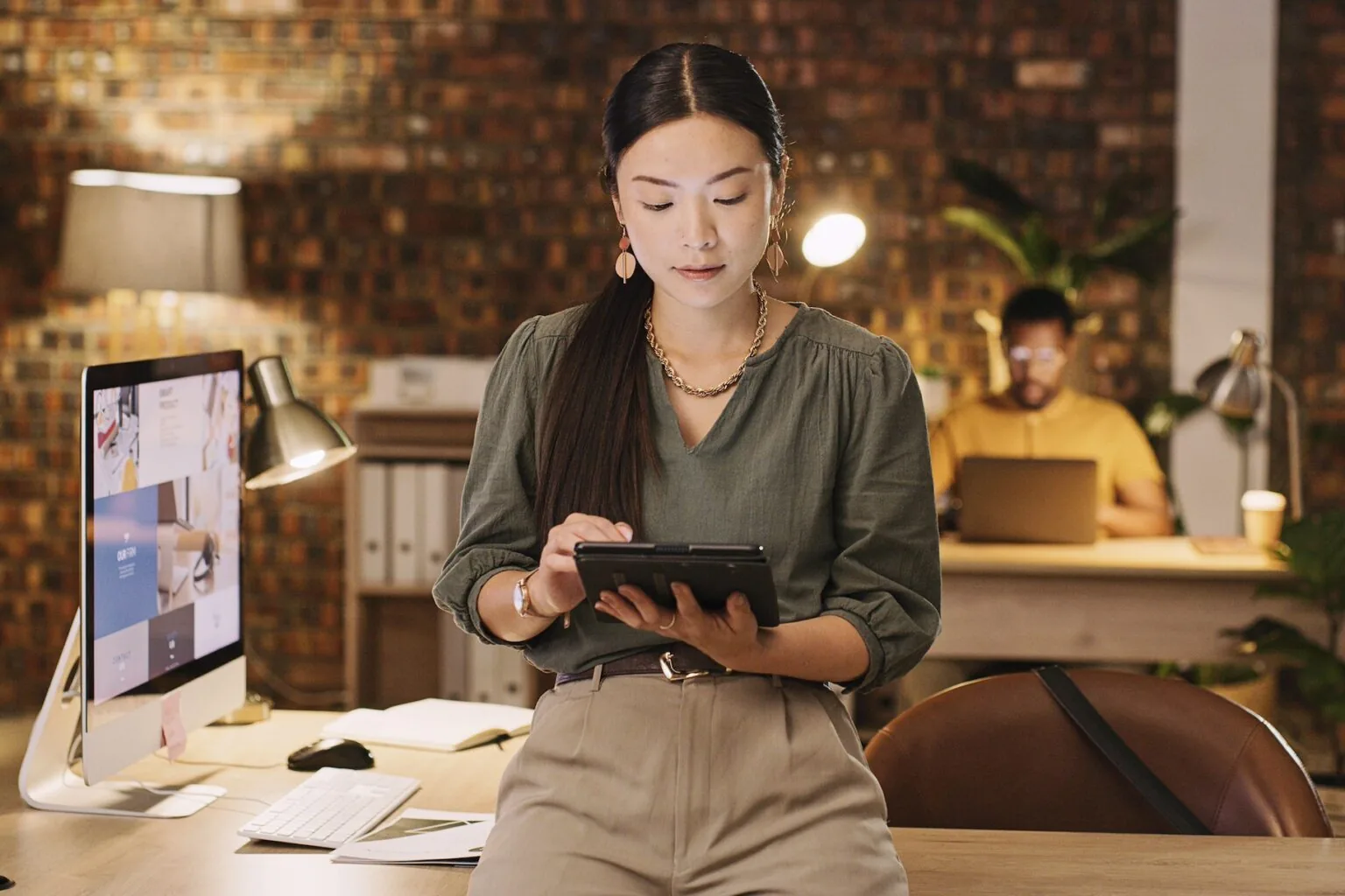 A woman wearing a green shirt, with khaki pants and gold jewelry accessories, is in an dimly lit office holding a tablet in an office leaning on the front of their 90 degree angle desk with an iMac computer to her left. Behind her is a man seated, wearing a yellow collared shirt, is seated behind his desk, in-front of a laptop.